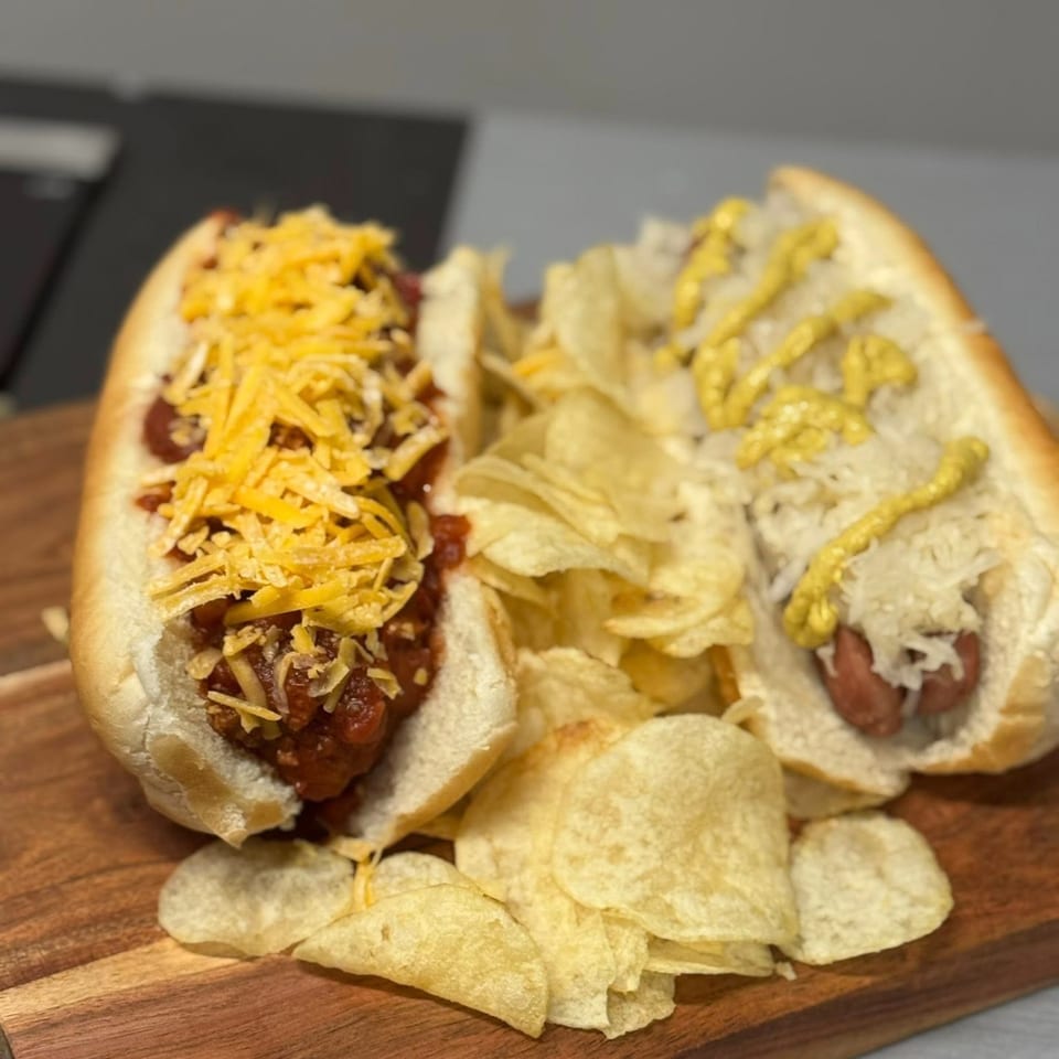 Two hot dogs, one with chili and cheese, the other with sauerkraut and mustard, served with chips.