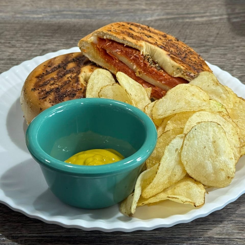 A grilled panini sandwich with salami and cheese, served with potato chips and a side of mustard.