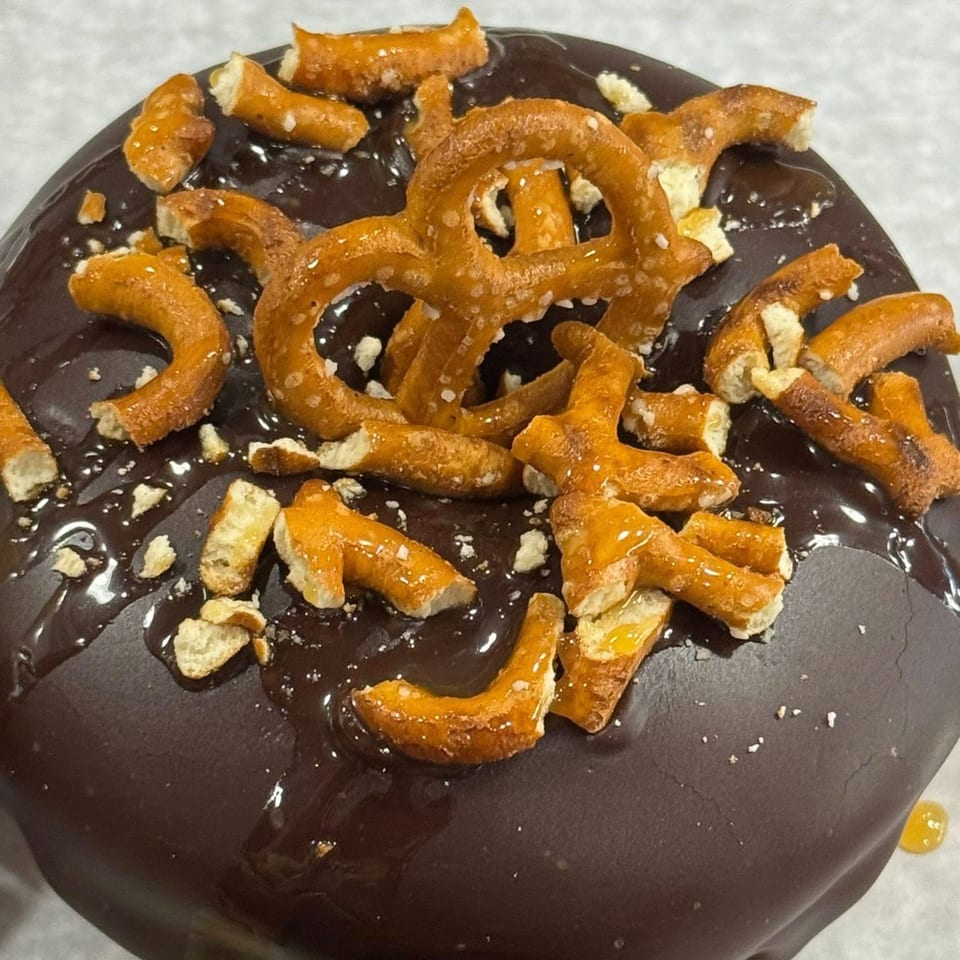 A close-up of a chocolate-covered donut topped with pretzel pieces and a drizzle of caramel.
